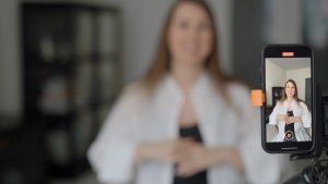A young white Woman applying to drama school in the UK. The woman is wearing a white shirt over a black t-shirt and is out of focus whilst a camera facing her on a tripod is in focus